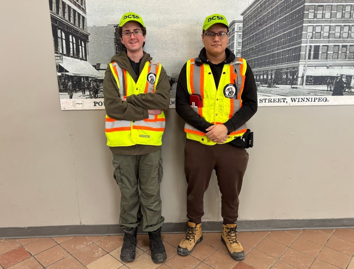 two-helpers-jack-ducak-and-seth-j-franklin Two young people wearing reflective vests, with their arms held in front of them standing in front of an old image of downtown Winnipeg.