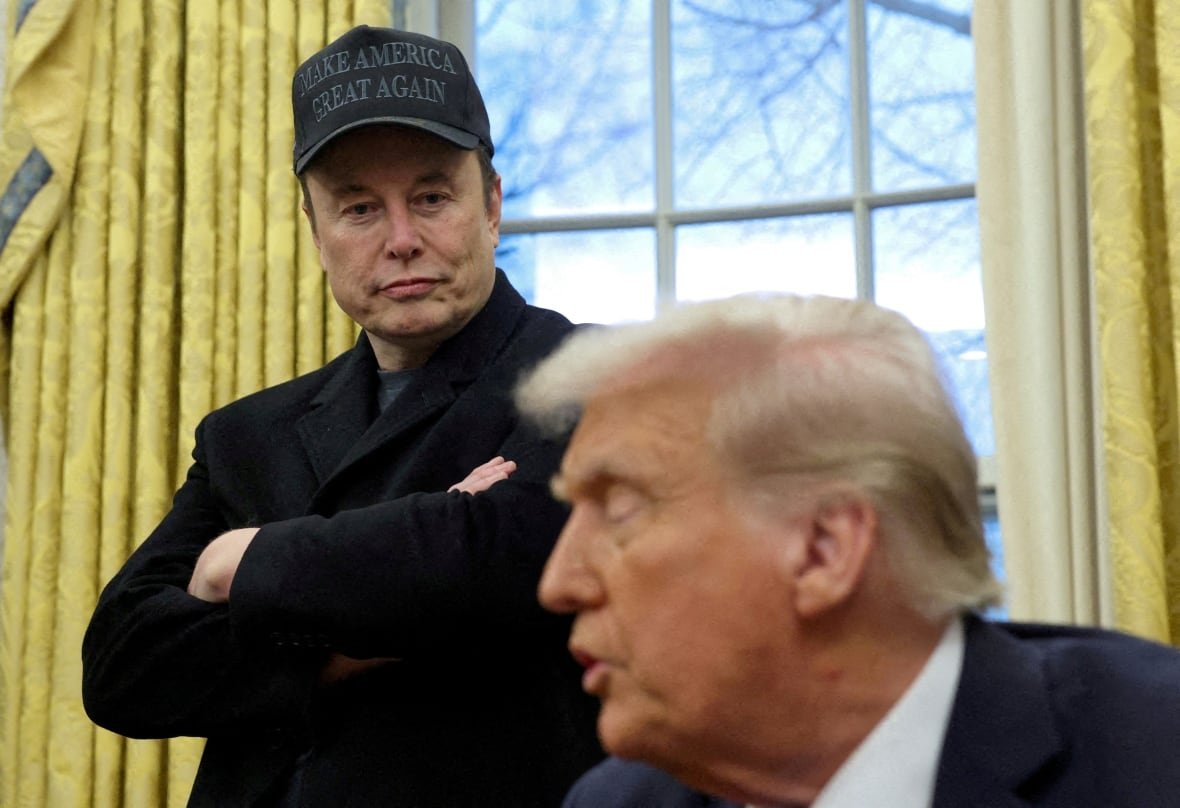 usa-trump-workers-cuts A man in a dark baseball hat and blazer stands next to a blond haired man who is seated.