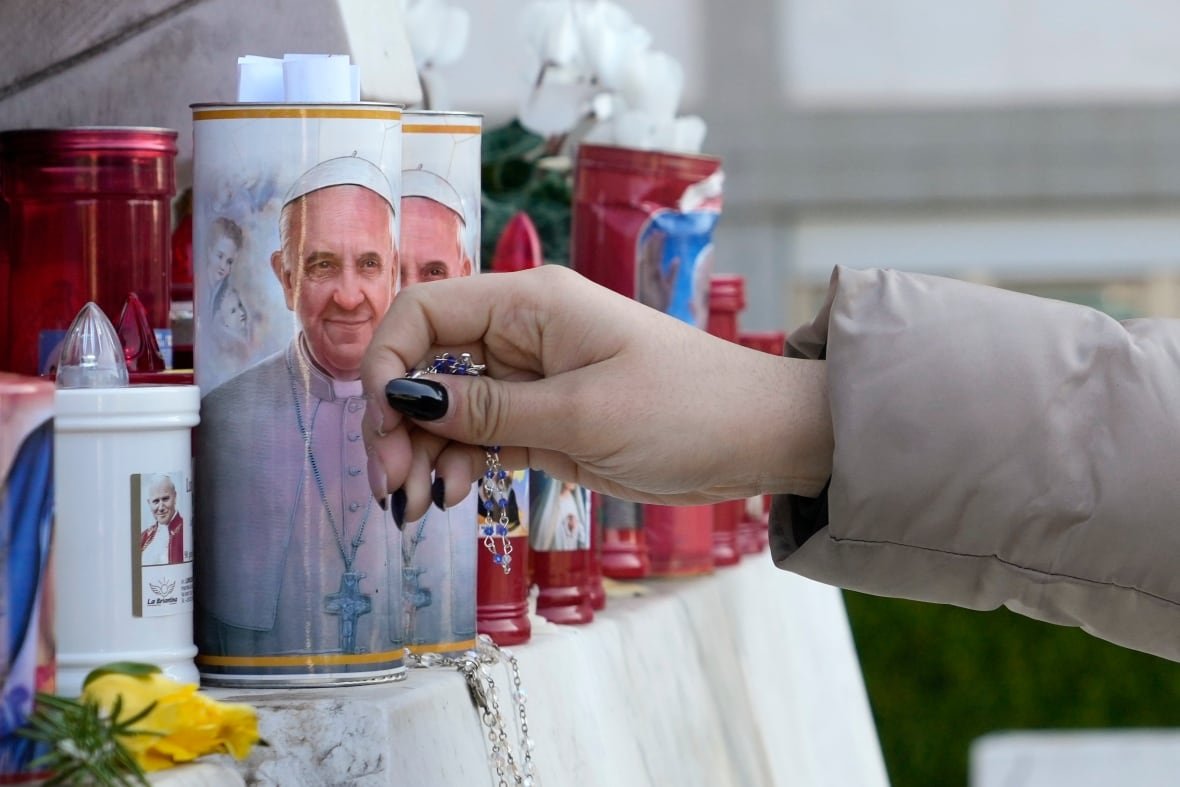 A person lays a rosary near candles adorned with a person's portrait.