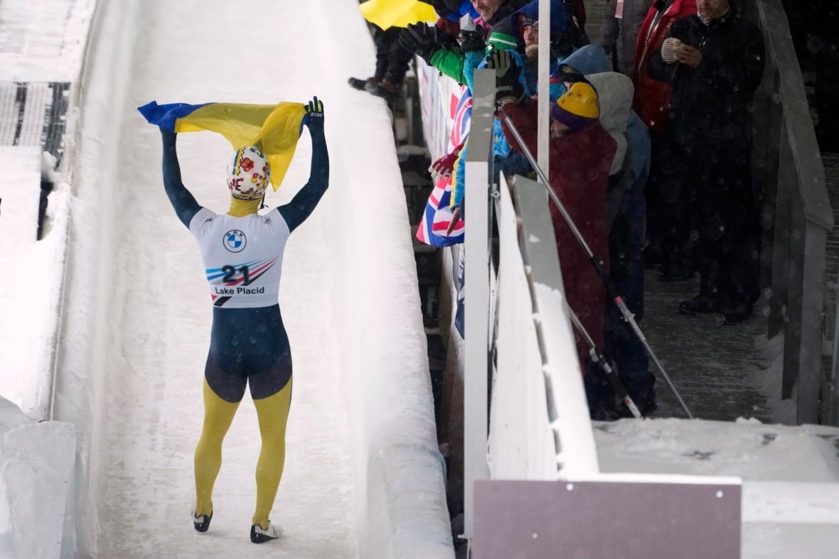 A male skeleton racer waves a Ukrainian flag as he stands on the track.