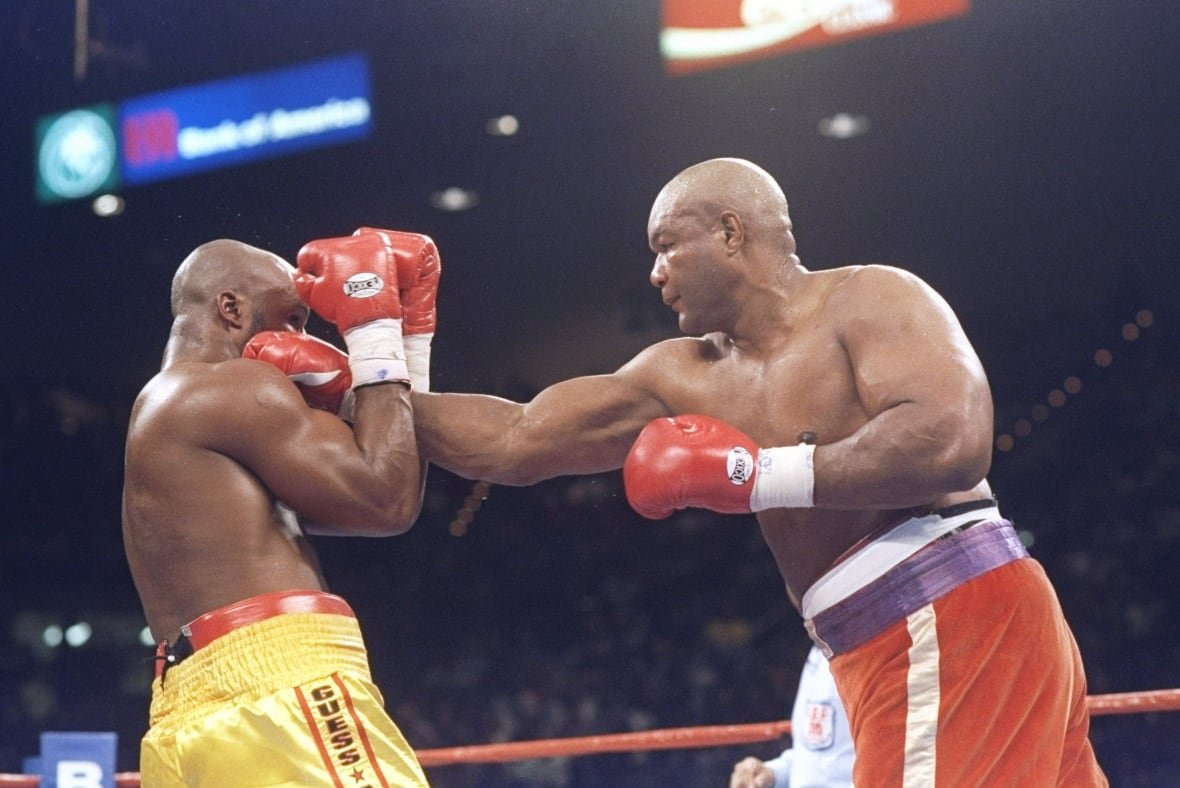 Two large, dark complected bald men are shown in a boxing ring. The fighter with red and blue trunks throws his right glove between the guard of the figther in yellow shorts.