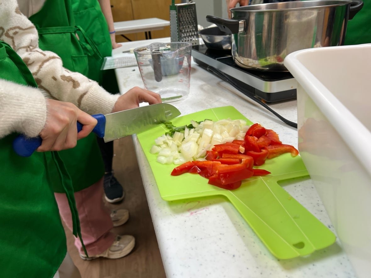 ecocooks-climate-friendly-cooking-1 Close-up image of a child's hands as they cut scallions next to a pile of chopped white onions and red bell pepper on a bright green cutting board.