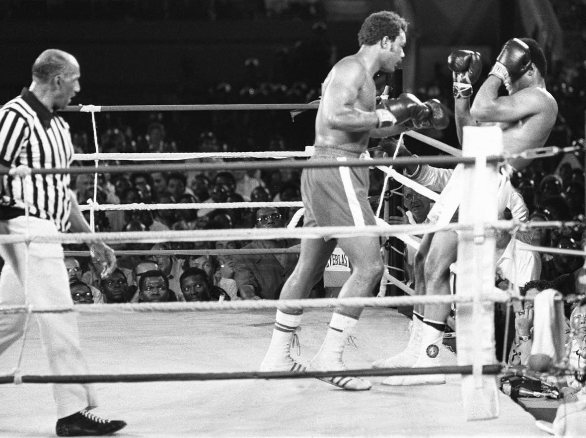 A black and white photo shows a boxing referee in the far left as two men in boxing gloves spar in the ring at right.