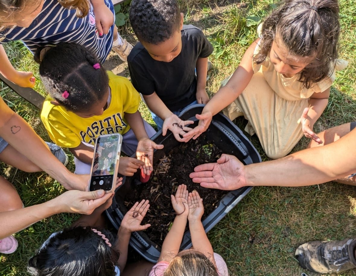 green-thumbs-growing-kids-1 A group of younger kids are seen from above outdoors, with their hands outstretched over a large bin full of soil and holding worms in their palms.