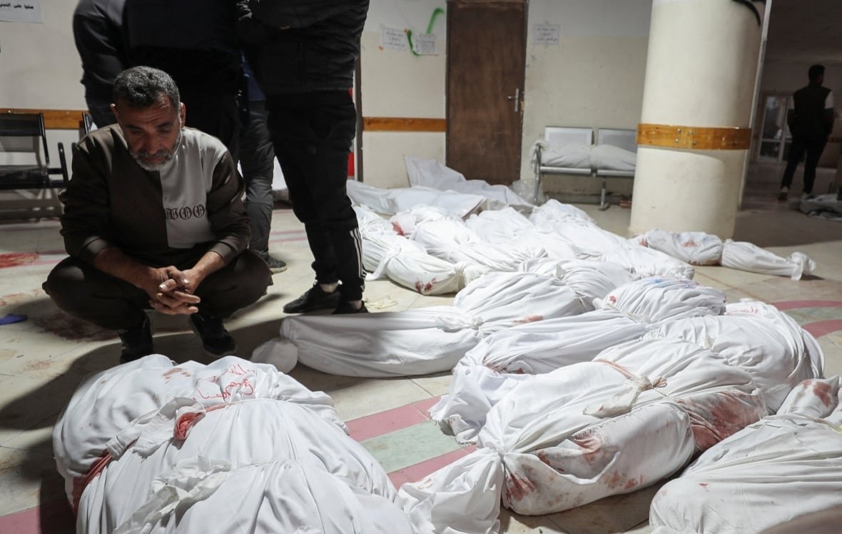 israel-palestinians-gaza-3 A man squats in a hallway filled with bodies wrapped in white shroud.