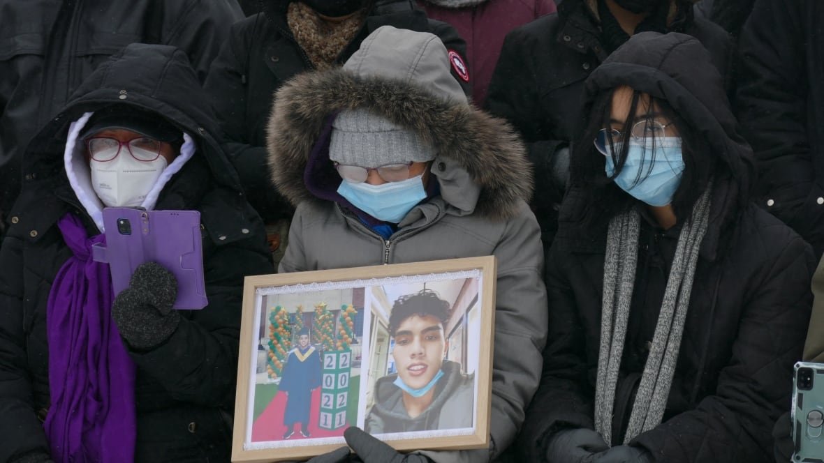 john-lloyd-barrion-vigil People wearing face masks and parkas stand together at a vigil. One holds two photos of a teenager in a frame.