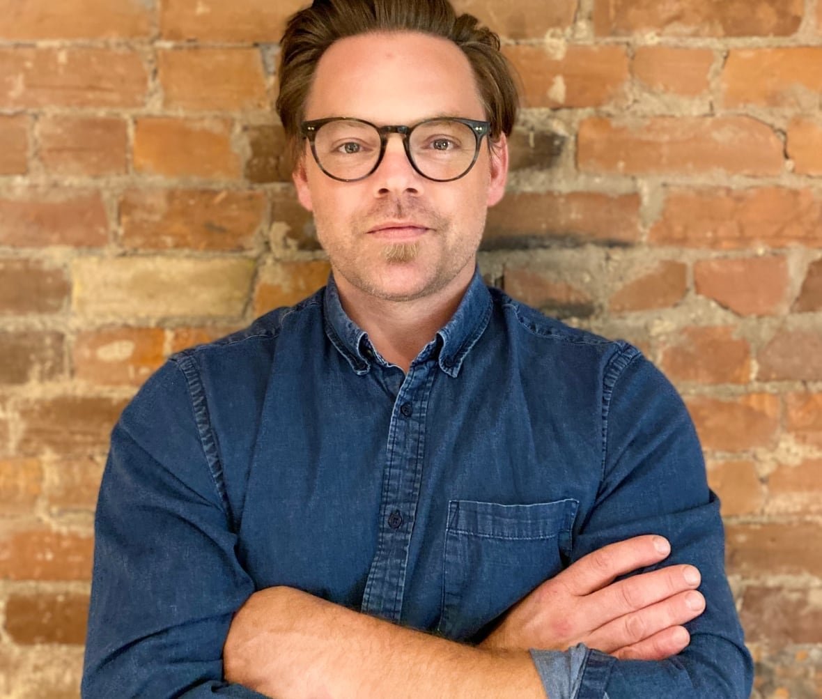 michael-classens A man in glasses and dark blue denim shirt stands against a brick wall with a neutral expression and his arms crossed.