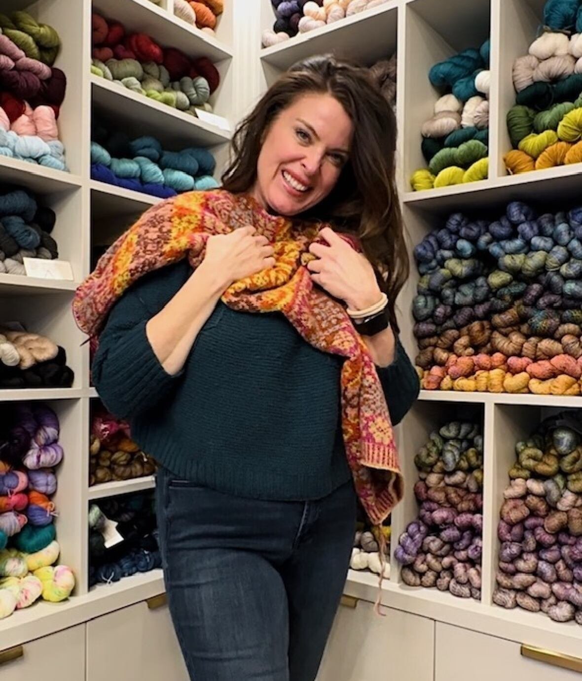 michele-bowland-jpeg A woman standing in front of a wall of colourful yarn and smiling at the camera while holding up a shawl to her chest.