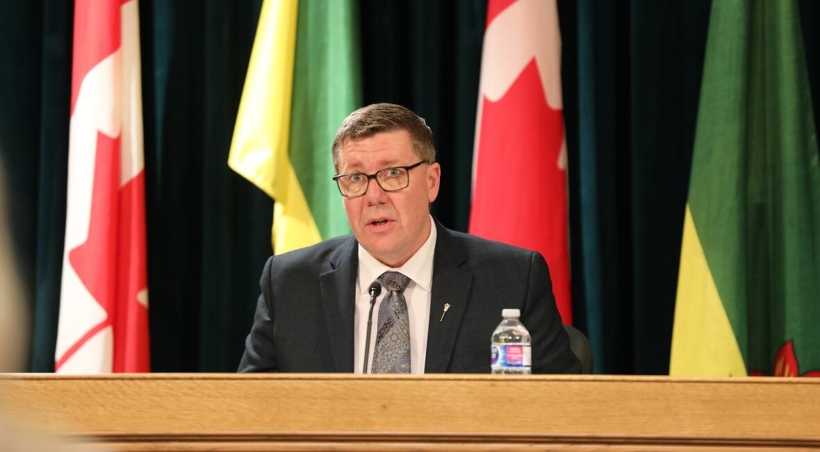 moe-budget-day A man in a suit with glasses speaks into a microphone at a desk. Behind him are several Saskatchewan and Canada flags.