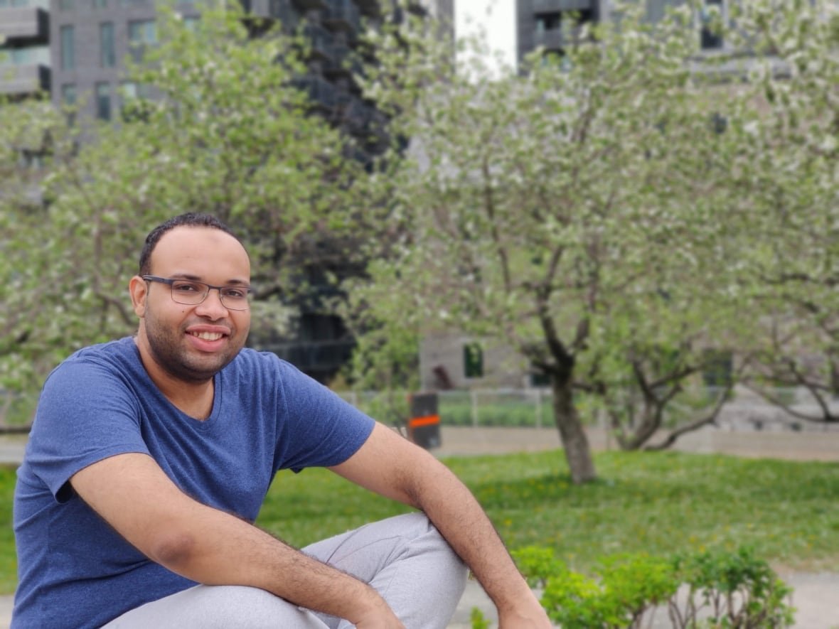 mohamed-hamid A man with a blue t shirt and glasses sits outside, smiling to camera.