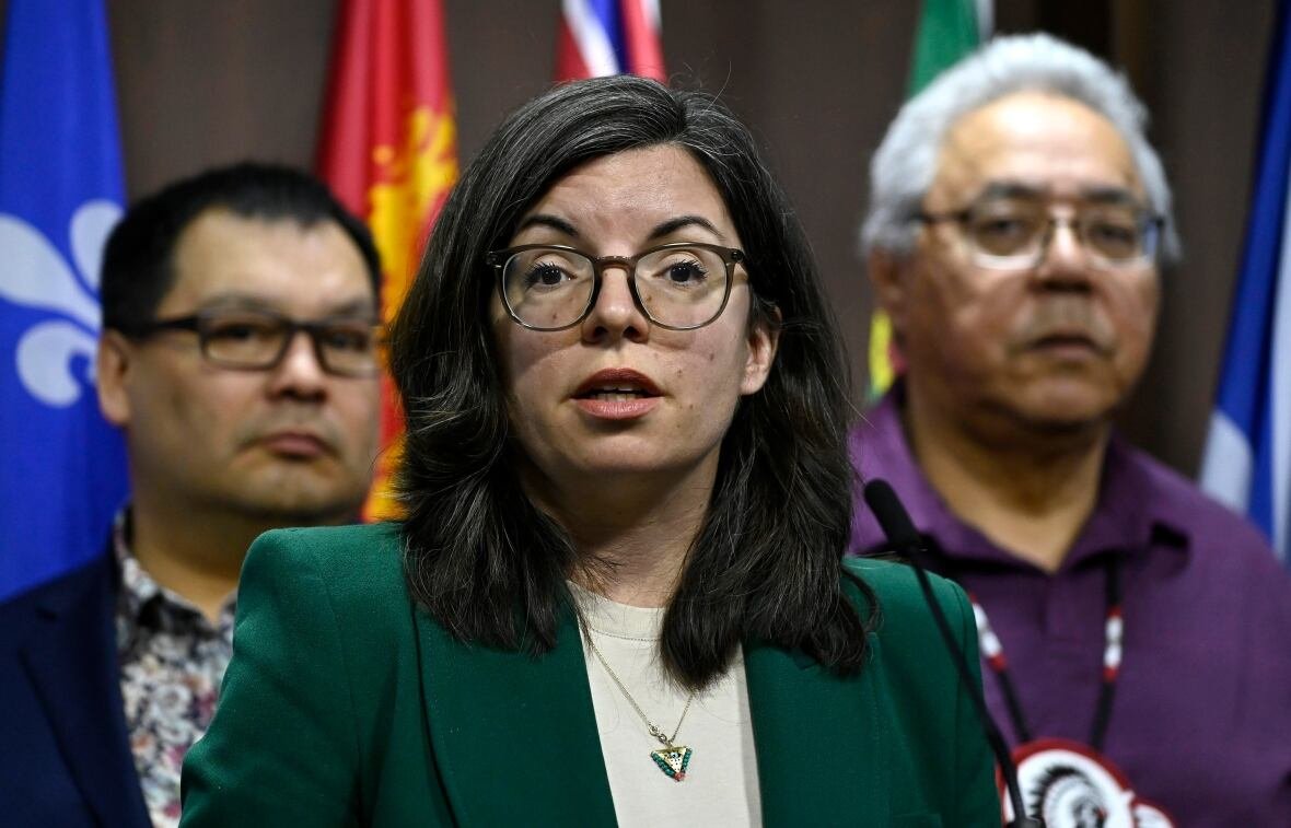 ndp-indigenous-health-20230403 A woman with glasses and a green blazer speaks into a microphone at a podium. Two people stand behind her.