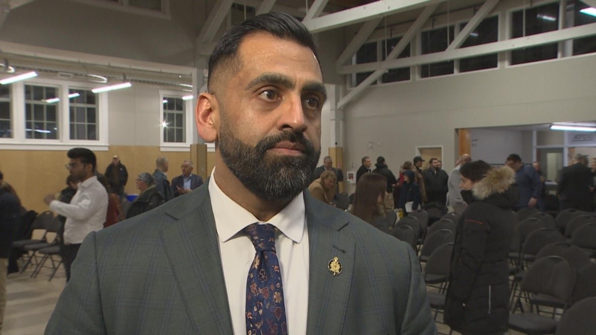 obby-khan A man in a grey suit with a white shirt underneath and a flowery tie stands in a room with people milling about behind him.