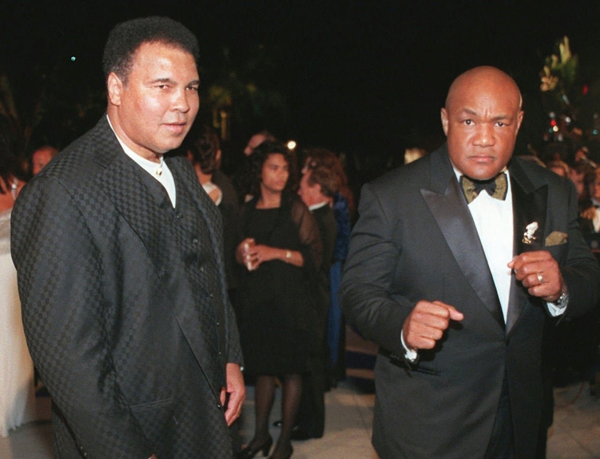 Two dark-complected, clean shaven men are shown next to each other wearing formal wear. One of the men, bald and in a tuxedo, strikes a boxing pose.