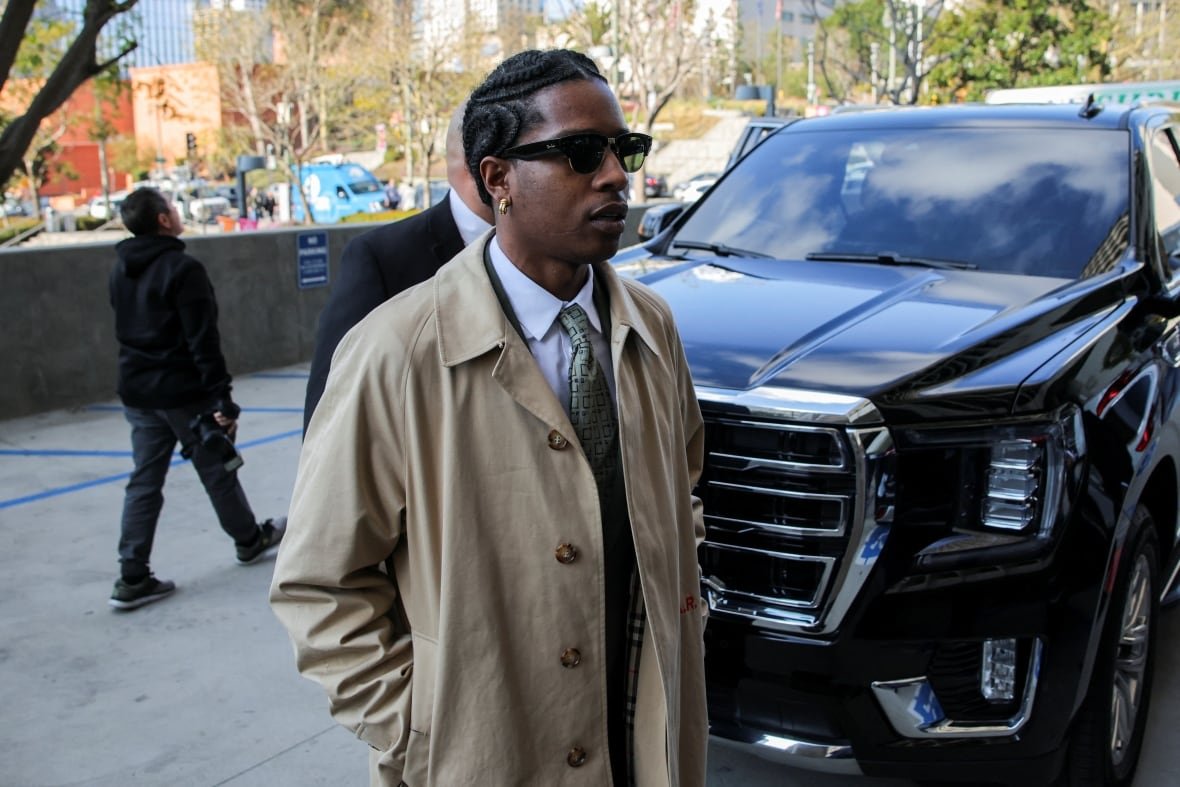 people-asap-rocky-trial A man with dark sunglasses and a beige trenchcoat over a suit and tie is seen in front of a black SUV.