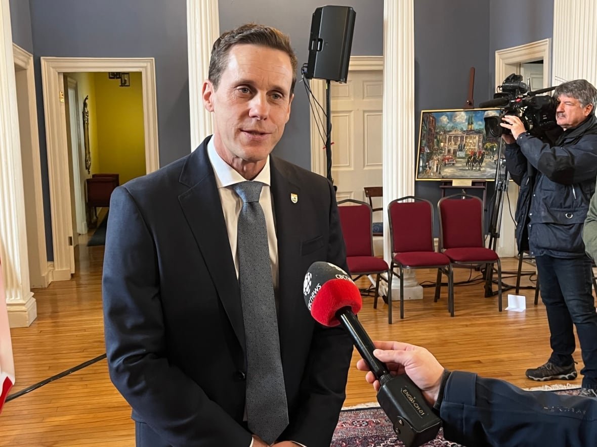 rob-lantz A man in a dark blue suit and tie answers questions from a reporter holding a microphone.