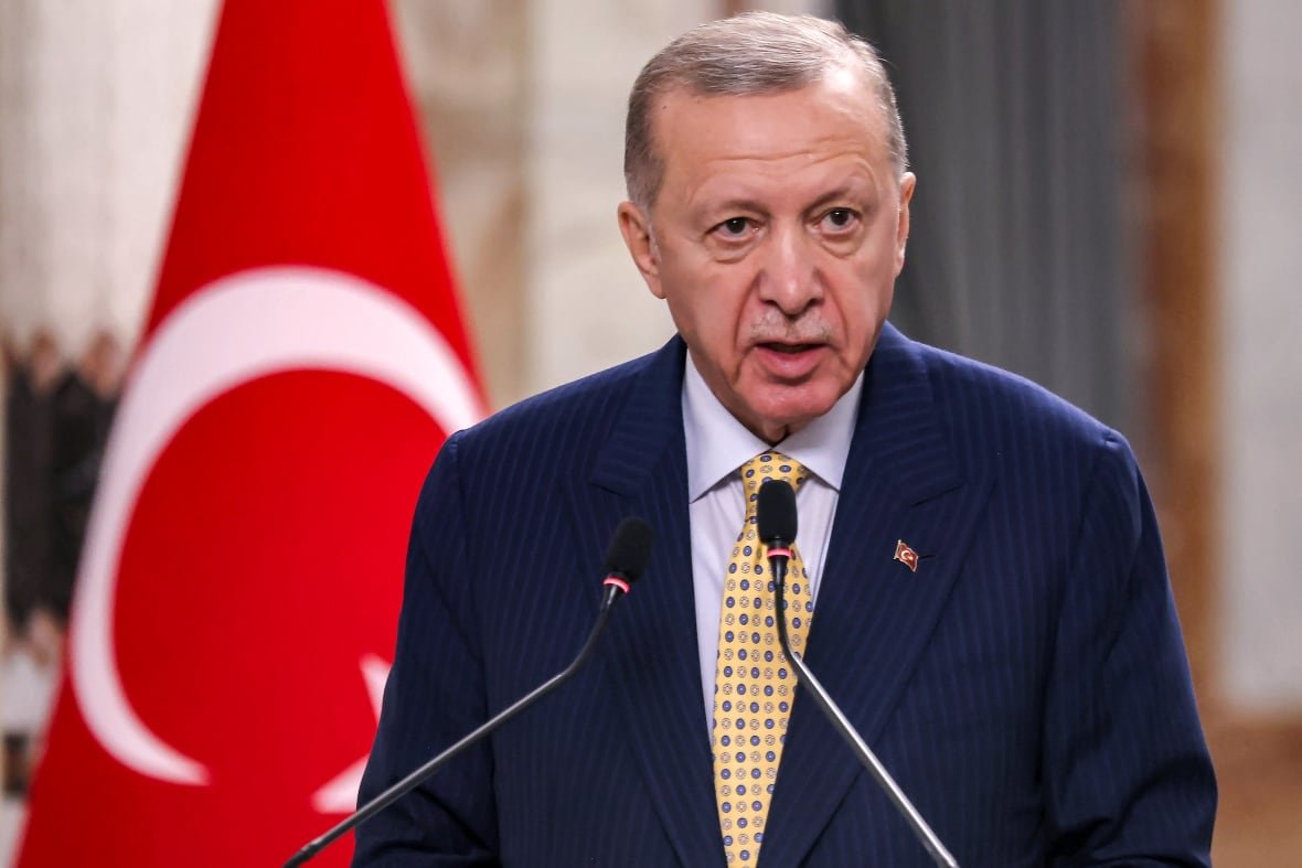 turkey-israel-trade An older man with a mustache wearing a suit and tie speaks into a microphone with the red flag of Turkey shown in the background.