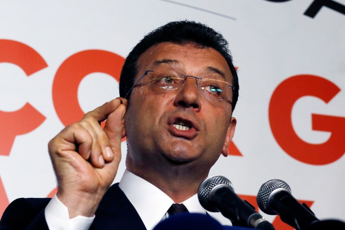 turkey-istanbul-election A man in a suit wearing glasses speaks in front of two microphones and gestures with his right hand.