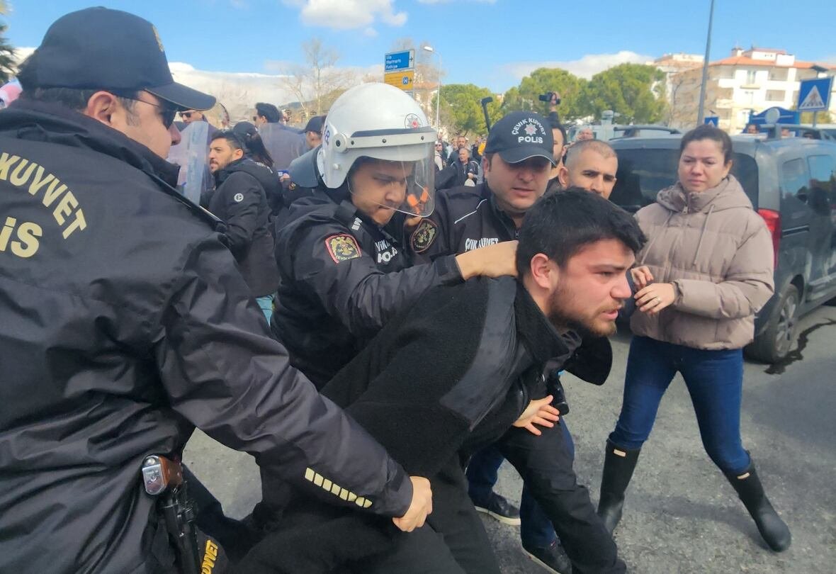 turkey-opposition-detention-mayor A dark haired man with a beard is grabbed by the back of the neck by a police officer in a helmet and riot shield, as other police and onlookers are shown.