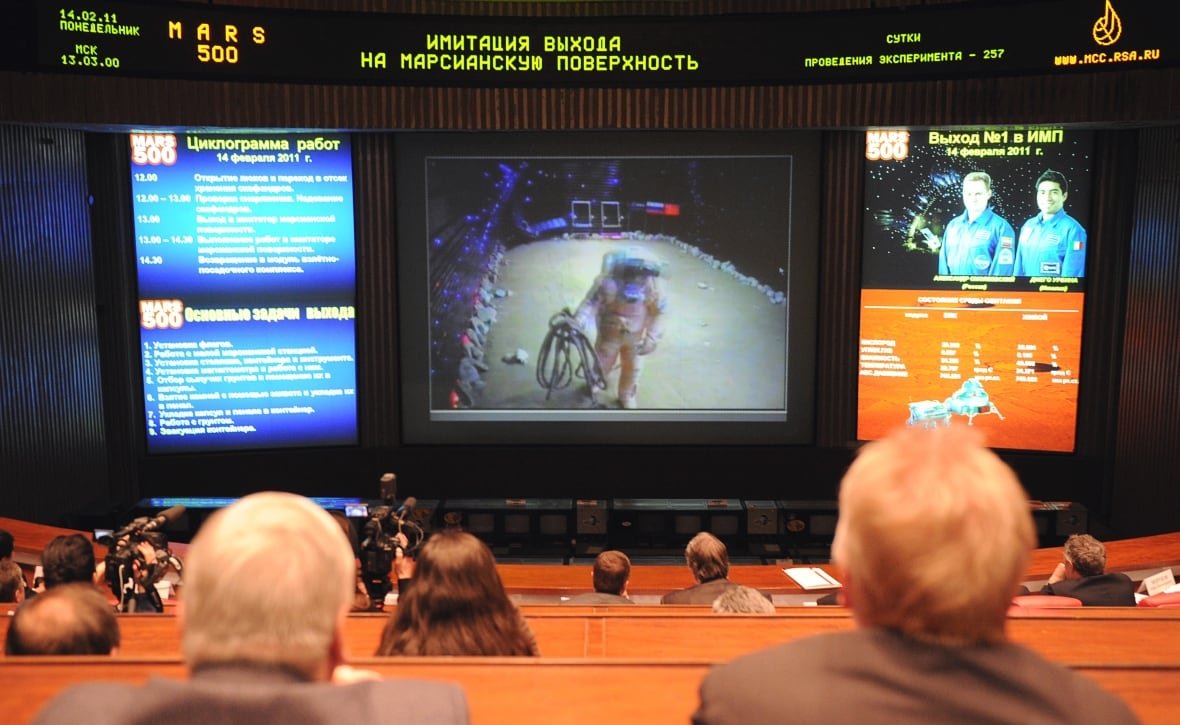 109100068 The backs of people's heads as they sit in a gallery watching people in spacesuits completing tasks on a large screen.