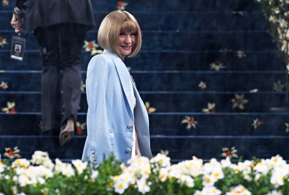 anna-wintour-met-gala-may-2025 A woman in a light blue coat poses for a photograph, her body turned to the right but her face toward the camera, as she walks behind a row of white flowers and in front of a blue carpeted staircase.