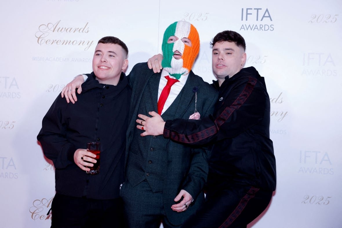 awamusic-kneecap-apology Three young men are shown hugging each other in a red carpet photo. One of them is wearing a ski mask over their head.