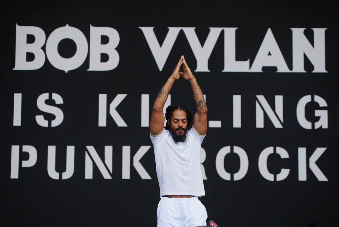 britain-glastonbury-music-festival-day-4-1 A man in a white t-shirt and shorts stands -- eyes closed, arms raised overhead and stands steepled -- in front of a sign onstage reading: 'Bob Vylan is killing punk rock.'