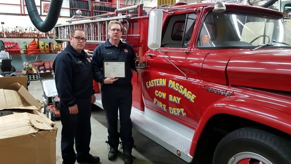 bruce-masales-2017-fire Two men in navy pants and sweaters with Halifax Fire crests on the chest stand beside a red firetruck in a garage. The man on the right has red hair and a moustache. He holds a certificate and small award.