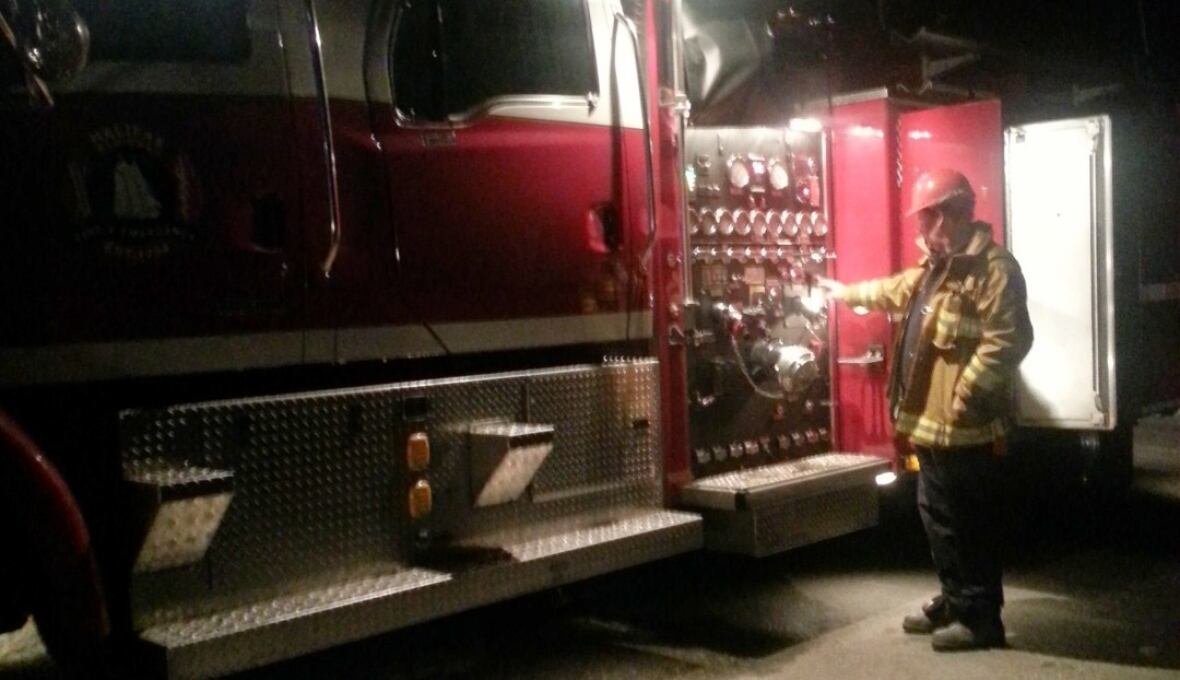 bruce-masales-fire-truck-2012 A man in firefighter gear, including a jacket with yellow stripes and red helmet, touches a red firetruck. The lighting is dark, with shadows around the man.
