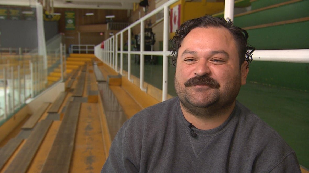cody-lightning A man with dark hair and a moustache, wearing a grey sweatshirt, sit in the stands in a hockey arena.