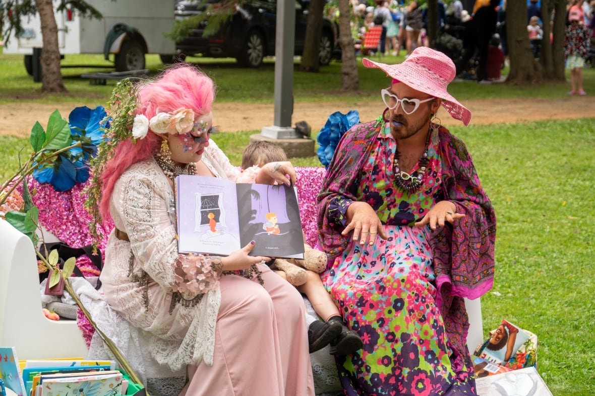 drag-queens Two people dressed in colourful clothing, mostly pink, sit outside and read a picture book.