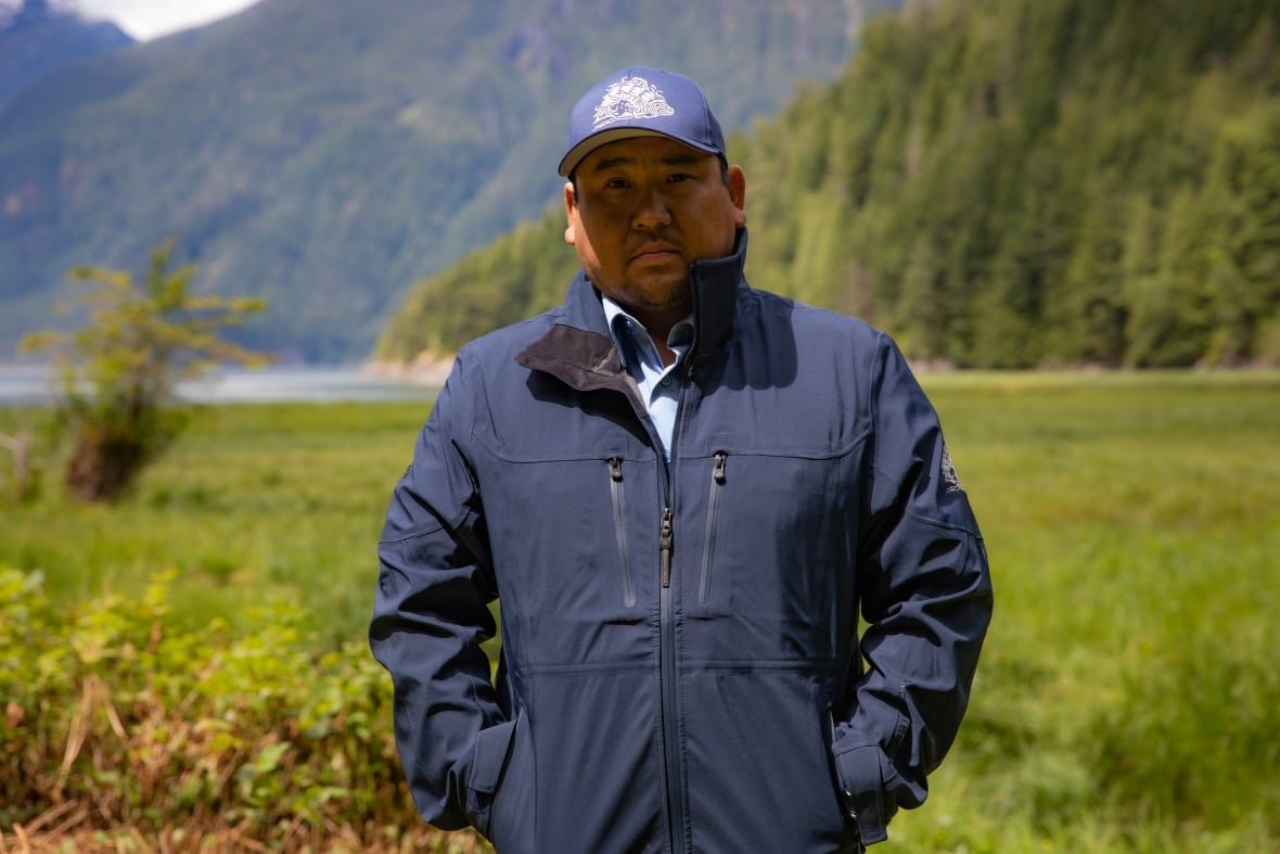 erik-blaney An Indigenous man stands outside, with water and a tree-covered mountain behind him.