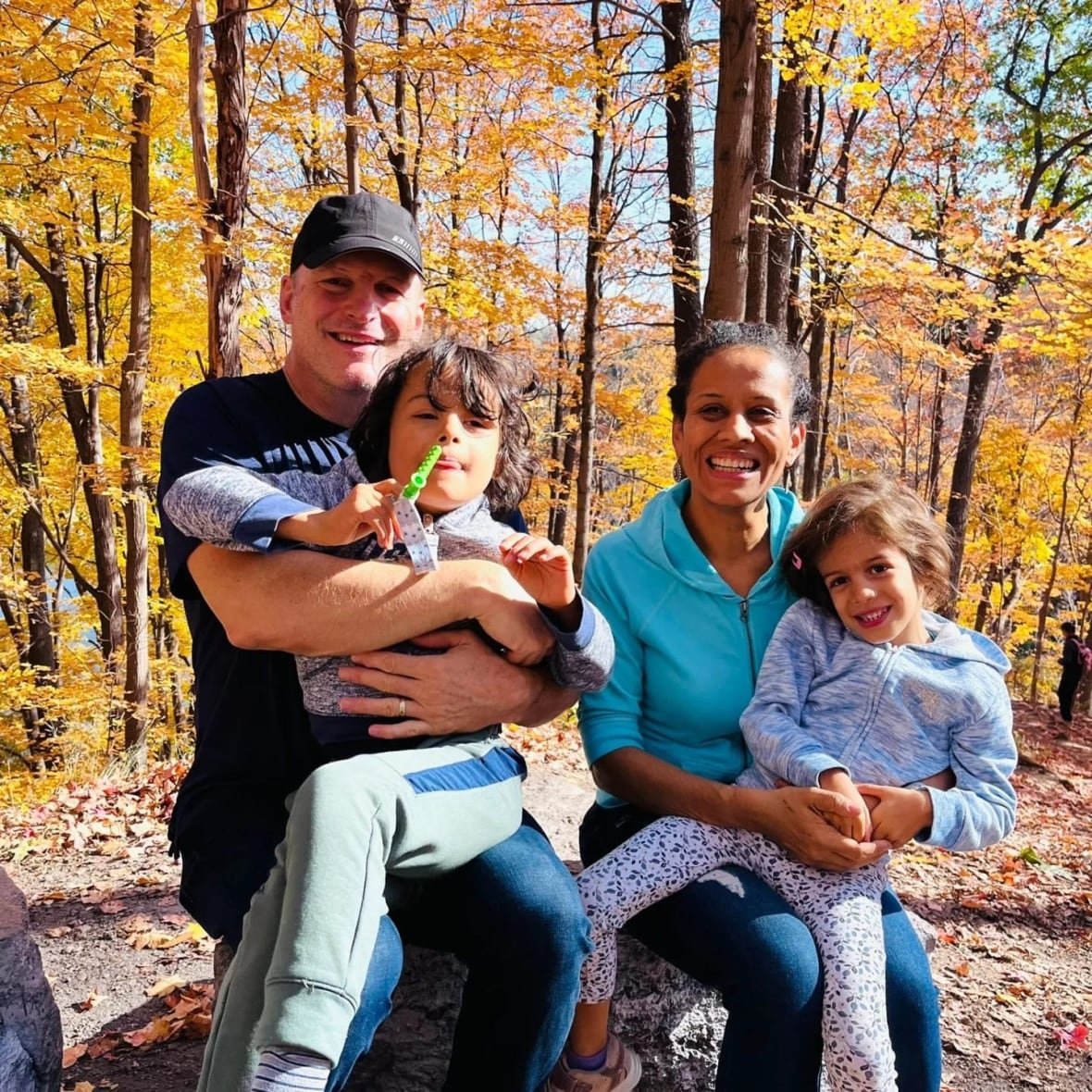 ferreira-legault-family A man (left) and a woman (right) are standing in a forest with fall-coloured leaves, holding an elementary aged boy and girl.