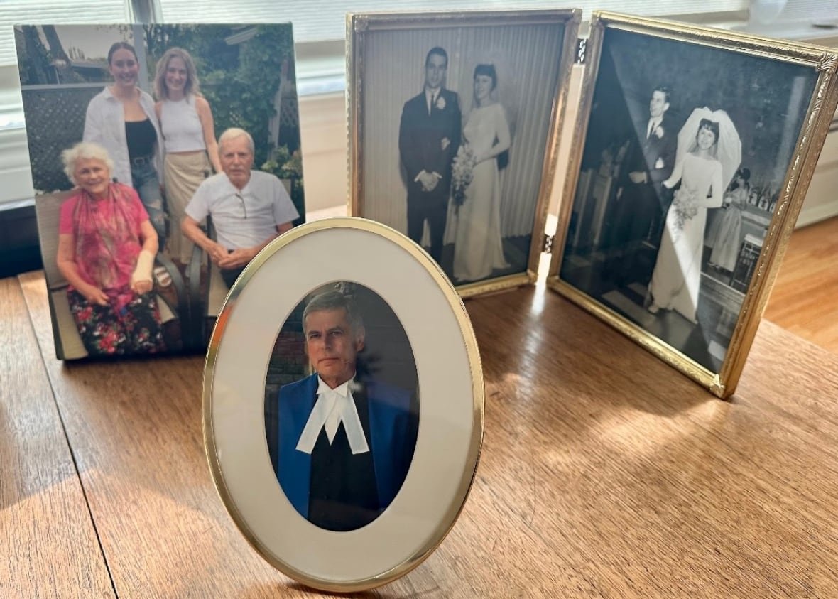 go-public-koshman-photos A series of framed family photos of the same man and woman at various points in their lives, including one image of the man in judge's robes, are displayed on a wooden table.