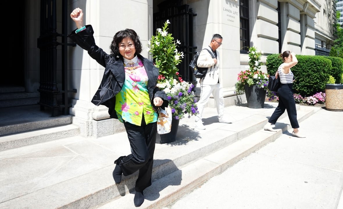 hudsons-bay-ccaa-20250623-2 A woman with a black bob, black jacket, floral shirt and black pants pumps a fist in the air, standing on the steps of a courthouse.