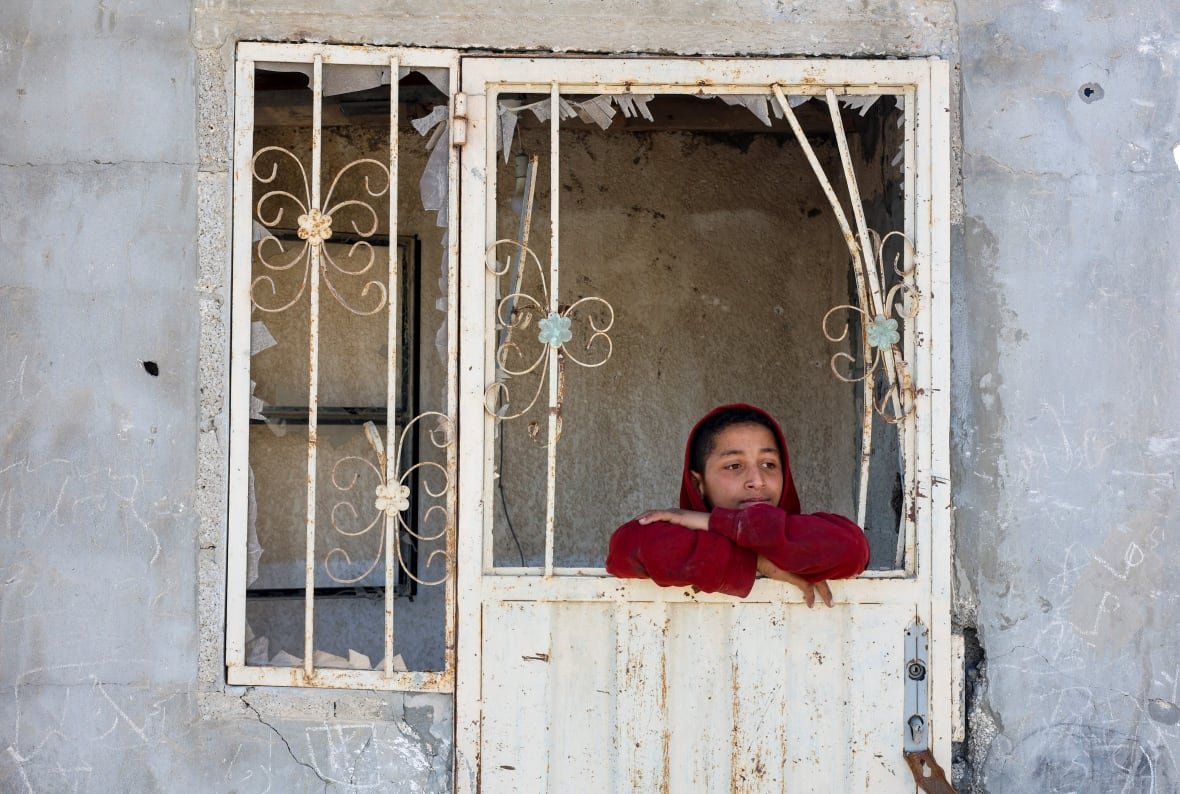 israel-palestinians-gaza A young boy in a red hoodie leans his chin on his arms as he looks out the window of a white house damaged in an airstrike. The glass in the window and door has been shattered.