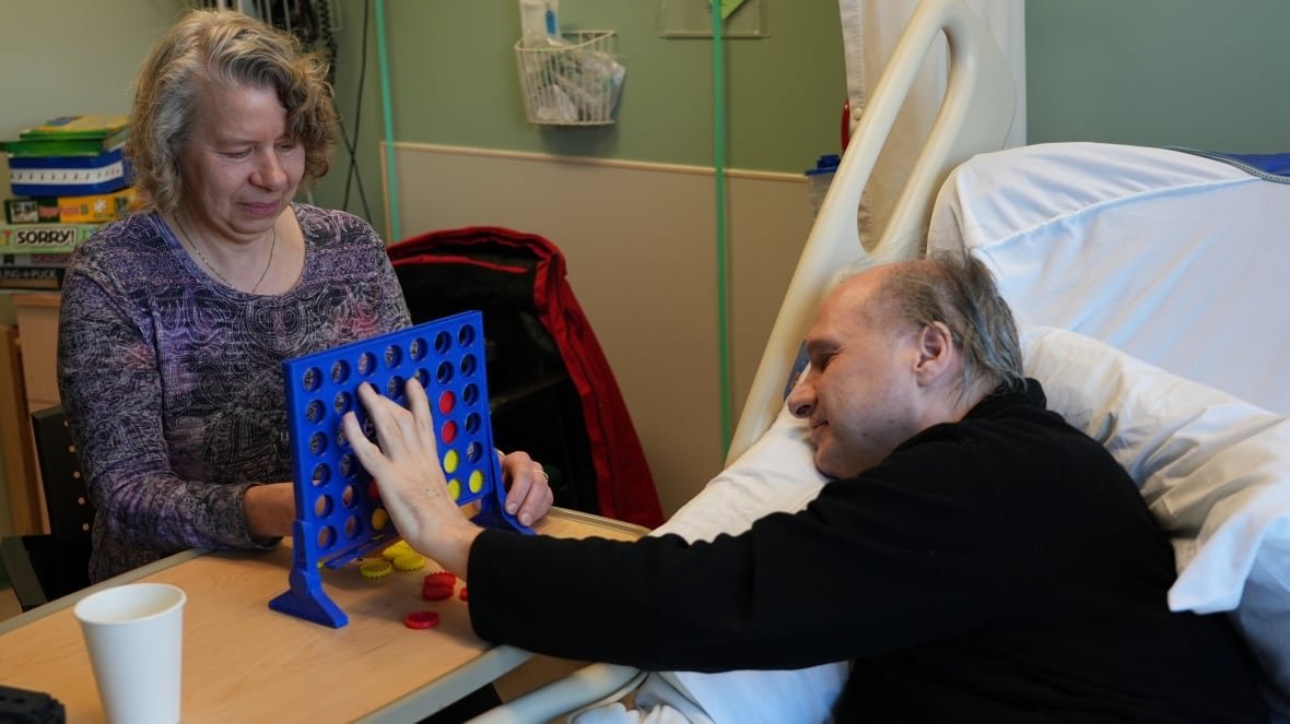 kareen-and-ken-rekowski-play-a-game-at-the-renfrew-victoria-hospital A woman and man play a game on a bedside table in a hospital room. The man is reaching from his hospital bed.