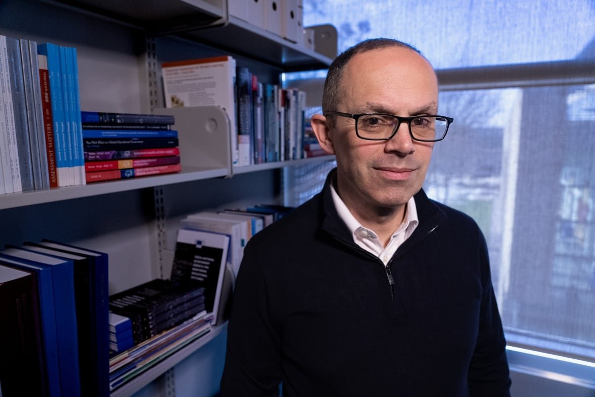 louis-volante A man in glasses, a white button down shirt and black sweater stands in his office, with shelves filled with books and a window seen behind him.