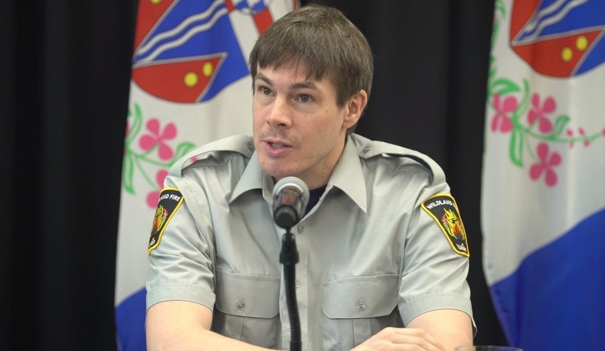michael-smith A man in a uniform shirt sits at a table speaking into a microphone.
