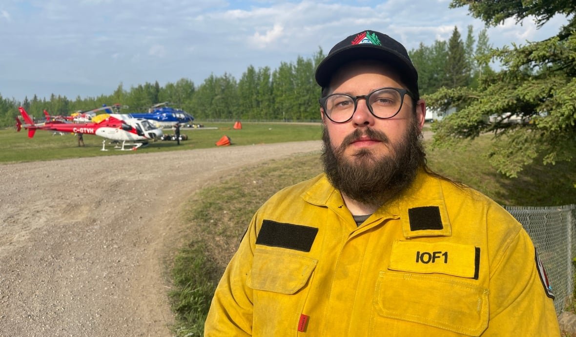 miuke-westwick-hay-river-kfn-fire A man in a yellow uniform with two helicopters on the ground in the distance.