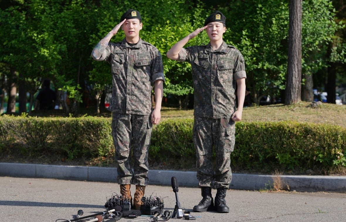 music-suga-bts-returns Two men in army fatigues stand on the pavement outside and salute. There are microphones on the ground at their feet.