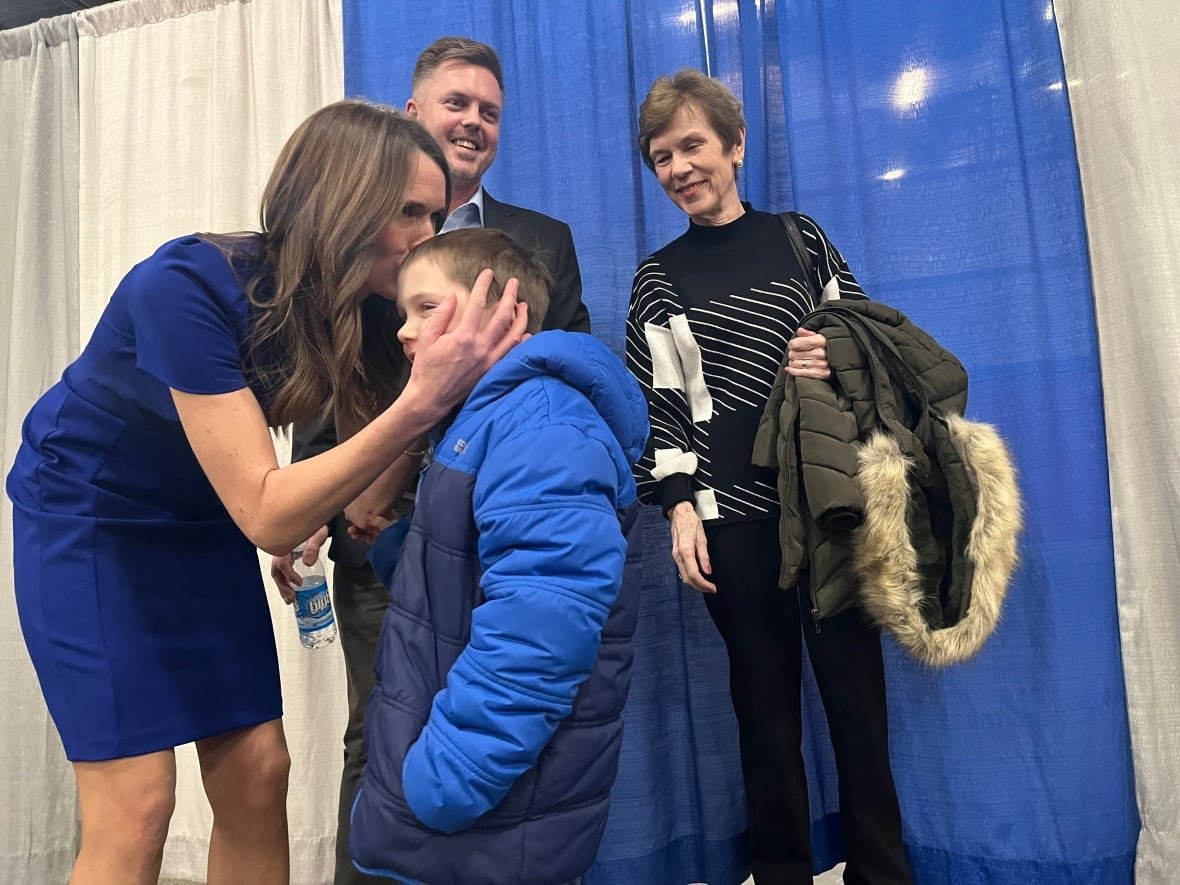 natalie-jameson A woman in a blue dress kisses her son on the head and two other people look on.