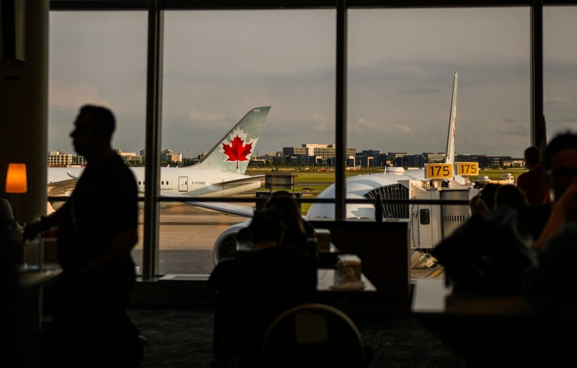 pearson-air-canada Travellers are seen in silhouette near a large window at an airport overlooking a plane set up for boarding.