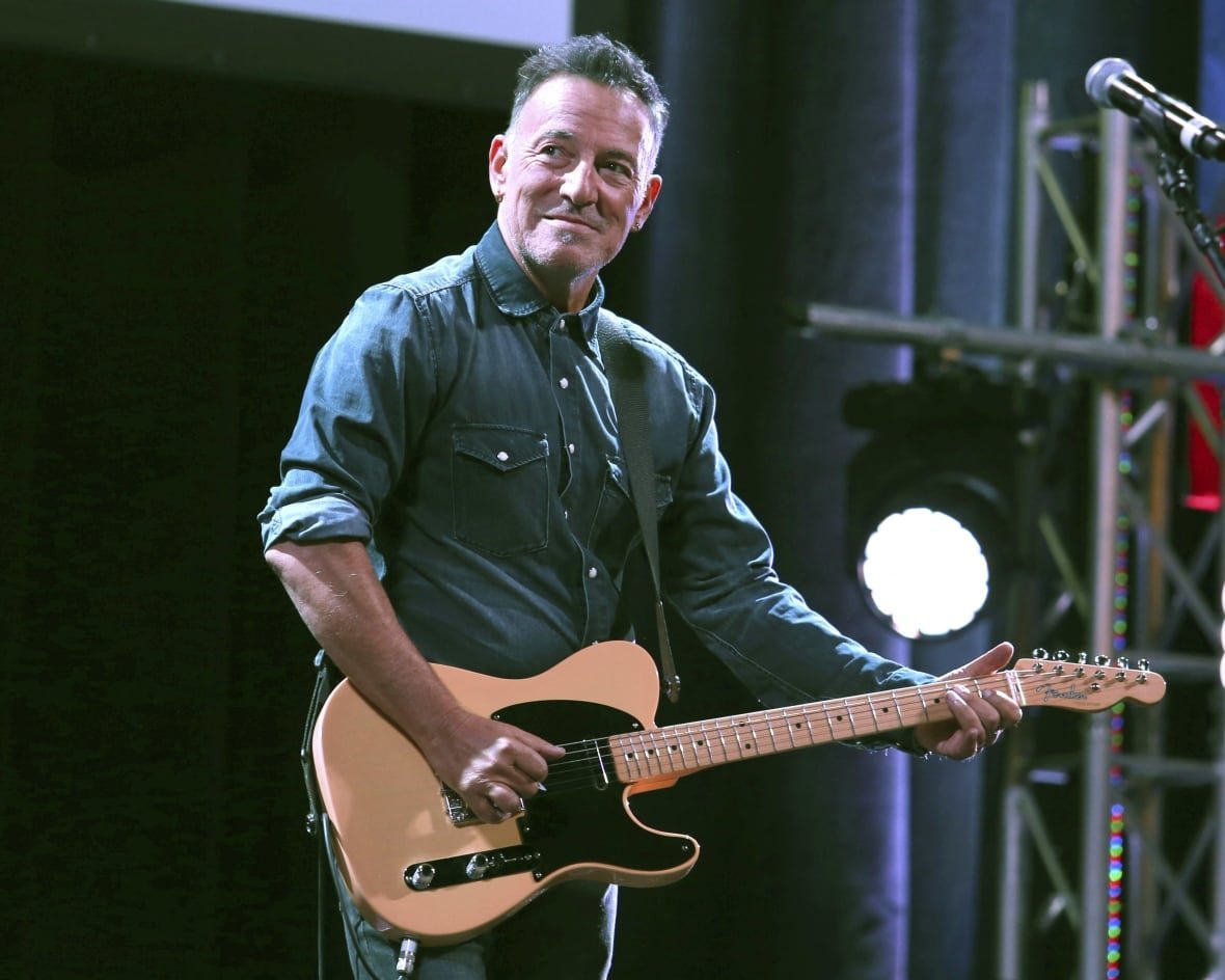 people-bruce-springsteen A man holding a guitar on stage looks to his right, smiling.