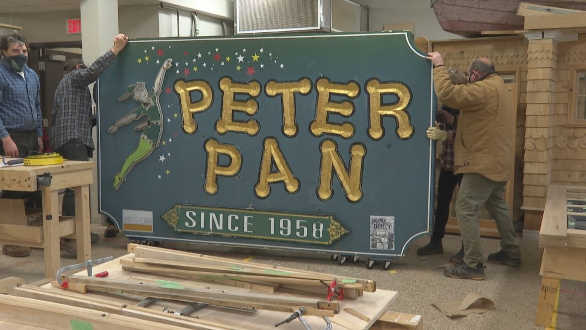 peter-pan-sign-arrives-at-holland-college-nov-2020 Several men position a large wooden sign that states 'Peter Pan: since 1958" in gold letters against a green background.