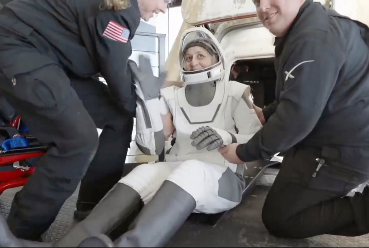 spacex-stuck-astronauts A woman in a spacesuit is sitting, she is being helped by two people in blue uniforms.