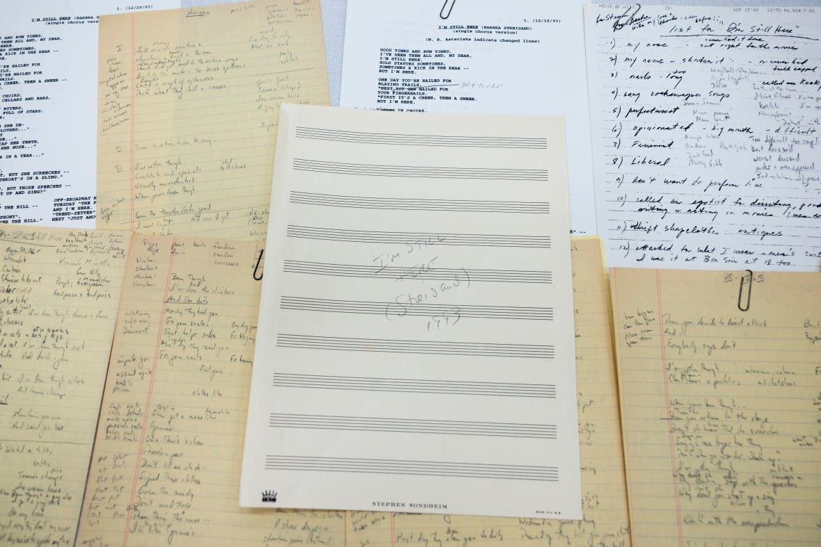 stephen-sondheim-library-of-congress Lined yellow paper with words scribbled on them and sheet music stacked on a table