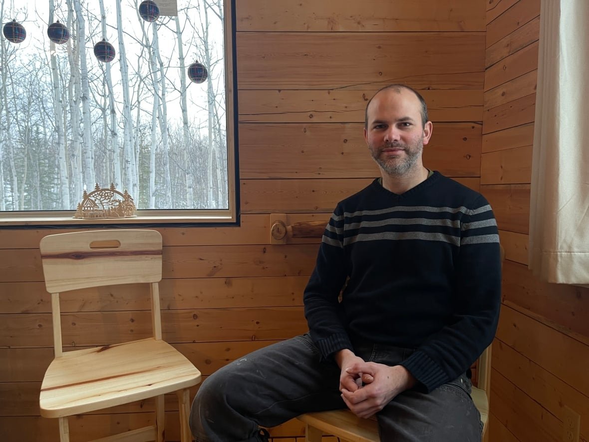 yukon-woodworker-ulrich-trachsel A man sits in a cabin beside a handmade chair. Outside the window is a aspen forest.