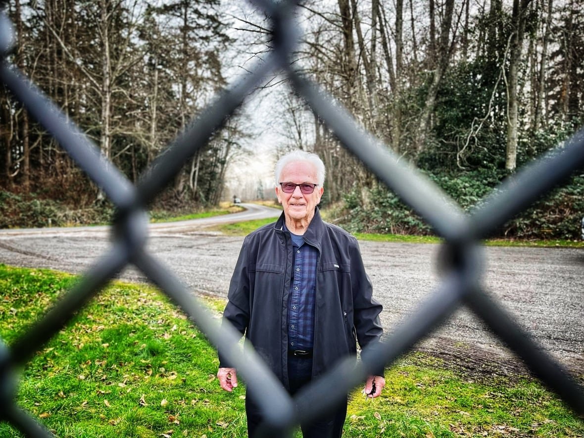 A man with white hair standing behind a black chain-link fence