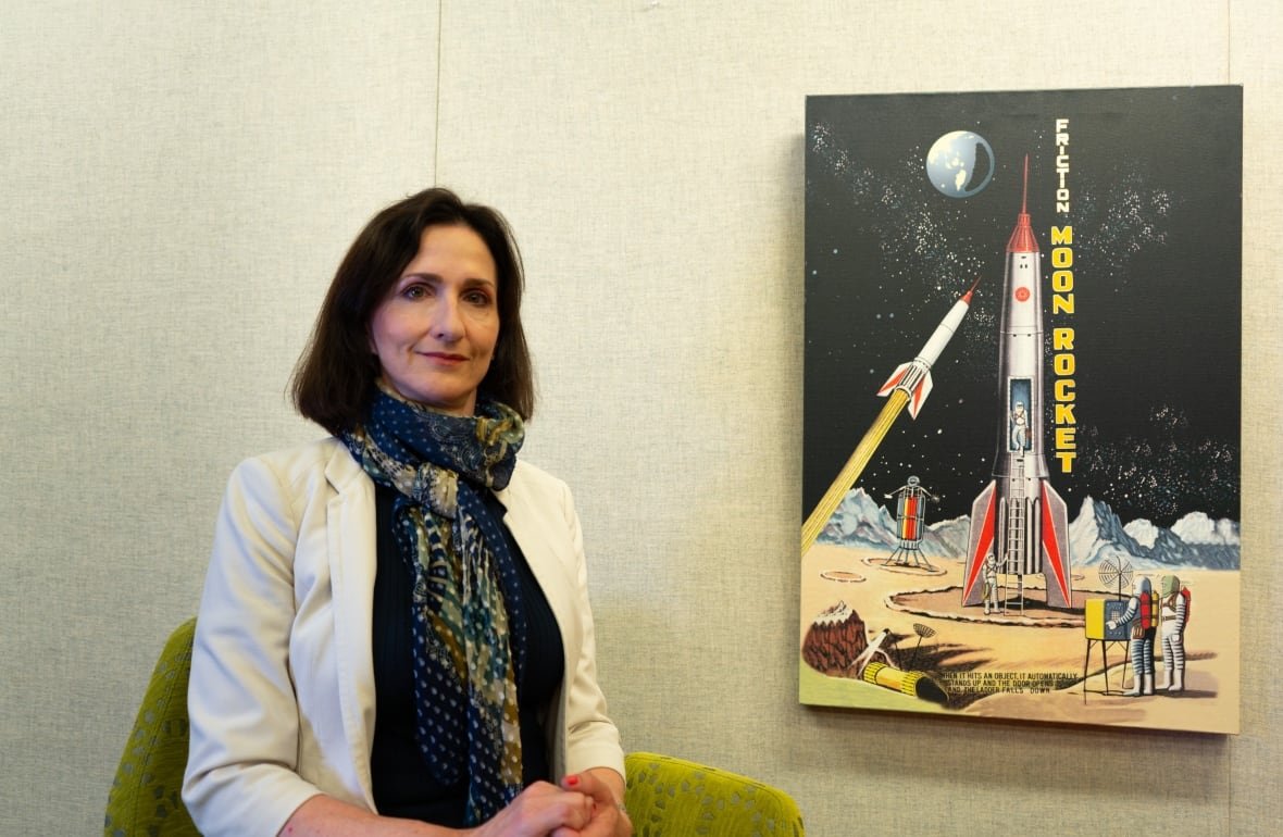 mit-astrophysicist-sara-seager A woman with shoulder-length hair wearing a scarf and white blazer poses for a photo next to an image of a rocket.