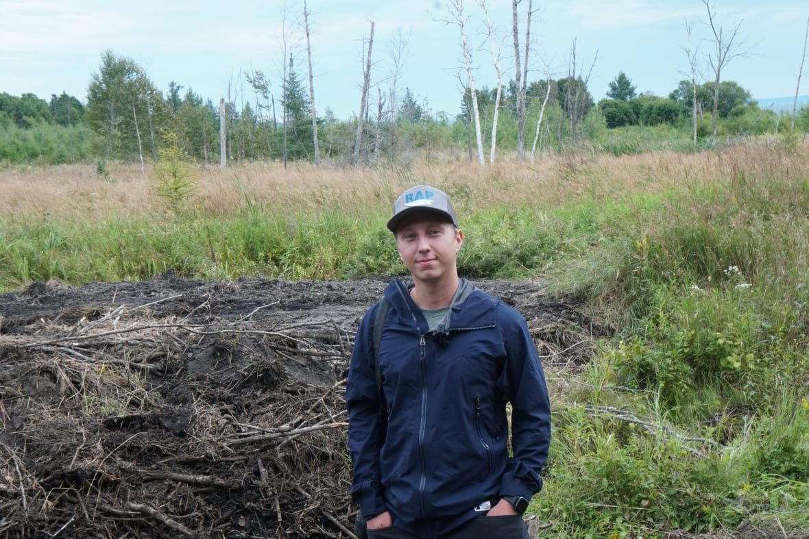 tim-hollinger A man in a blue jacket and grey hat stands in front of a pile of mud and a grass field